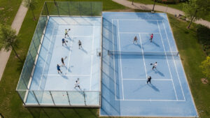 Vue aérienne de deux terrains voisins : un court de padel vitré et un court de tennis, avec des joueurs débutants en tenue sportive.